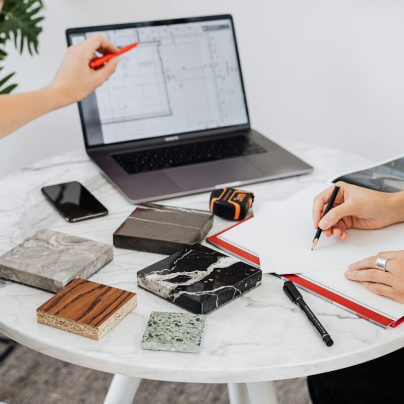 Two designers collaborate on a project with laptop and material samples on a marble table.